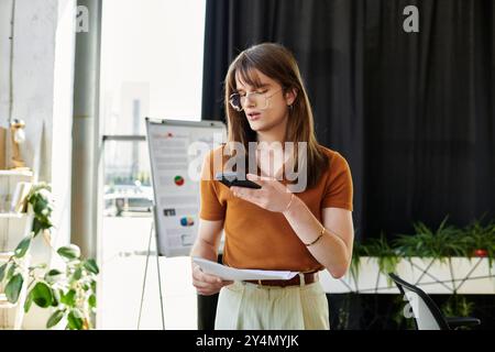 Una giovane persona non binaria esamina le note mentre utilizza uno smartphone in un ufficio luminoso. Foto Stock