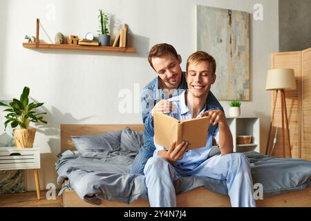 Una felice coppia gay condivide un delizioso momento mattutino in pigiama, leggendo e abbracciando a letto. Foto Stock