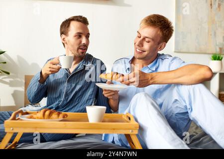 Una felice coppia gay si diverte a fare colazione nel pigiama, assaporando la compagnia degli altri e deliziose prelibatezze. Foto Stock