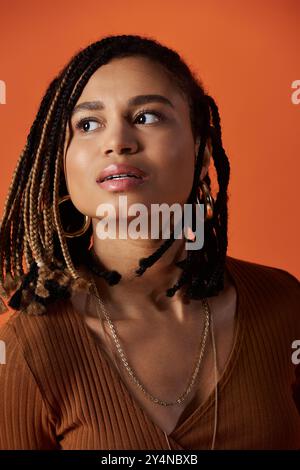 Una giovane donna sicura di sé con i capelli intrecciati sfoggia il suo look elegante in un'atmosfera calda e colorata da studio. Foto Stock