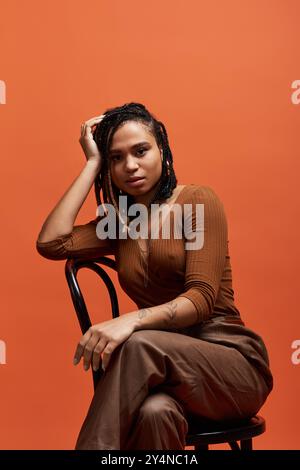 Una giovane donna afroamericana con i capelli intrecciati mostra il suo stile mentre posa su uno sfondo arancione. Foto Stock