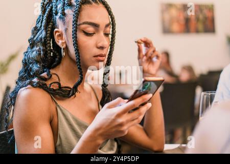 Giovane donna afroamericana elegante con capelli intrecciati che usa lo smartphone al ristorante. Stile di vita moderno, social media e tecnologia mobile con Foto Stock