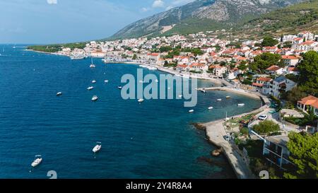 Annidate lungo la splendida costa dell'isola di Brac, le case vivaci punteggiano il paesaggio mentre le barche ondeggiano dolcemente nelle acque cristalline. Il caldo sole bagna il villaggio, creando un paradiso pittoresco. Foto Stock