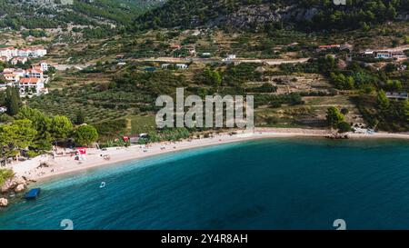 I visitatori si rilassano sotto il sole su un'incantevole spiaggia di Bol Brac Island, mentre le acque cristalline si infrangono dolcemente sulla riva. La vegetazione lussureggiante sale sulle colline che fanno da cornice a questa idilliaca fuga. Foto Stock