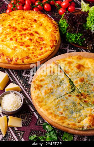 Due pizze al formaggio o khachapuri su tavole di legno accanto a ingredienti freschi su una tovaglia decorativa Foto Stock