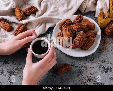 Mani che tengono una tazza di caffè accanto a un piatto di biscotti al cioccolato su un tavolo rustico Foto Stock