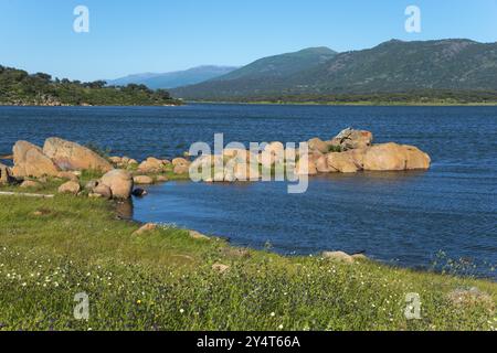 Paesaggio con lago calmo circondato da rocce e prati, montagne sullo sfondo sotto un cielo azzurro, il bacino idrico di Embalse de Plasencia, Embalse Foto Stock