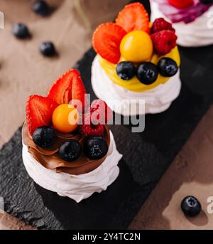 Tre mini pavlovas con panna montata e frutti di bosco freschi su un elegante tavolo da servizio in ardesia Foto Stock