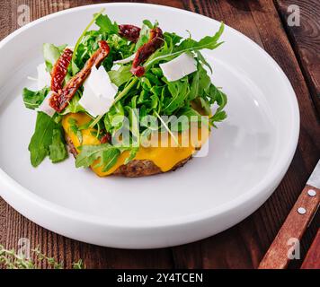 Delizioso cheeseburger con rucola e pomodori secchi su un tavolo rustico con contorni Foto Stock