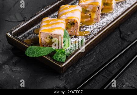 Eleganti sushi Rolls con kiwi e banana serviti su un piatto con un ramo sakura, su uno sfondo di pietra scura Foto Stock