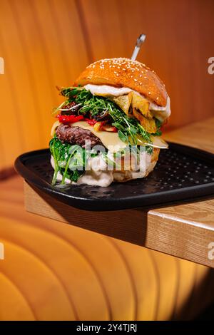 Hamburger artigianale su piatto nero con formaggio, verdure e pomodori secchi, in un ambiente accogliente Foto Stock