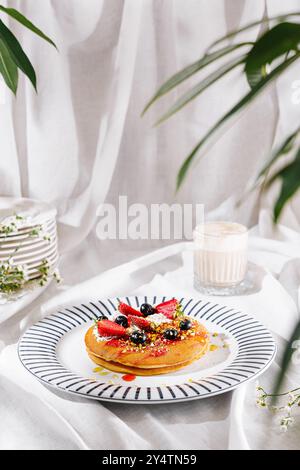 I pancake con panna montata, frutti di bosco e pistacchi si trovano su una tovaglia bianca vicino al cappuccino e alle foglie di palma Foto Stock