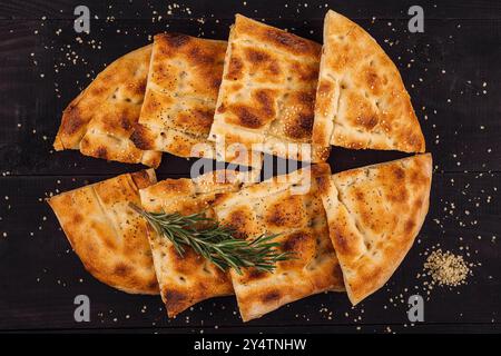 Pane turco pide appena sfornato, tagliato e condito con semi di sesamo e rosmarino, esposto su un tavolo di legno scuro Foto Stock