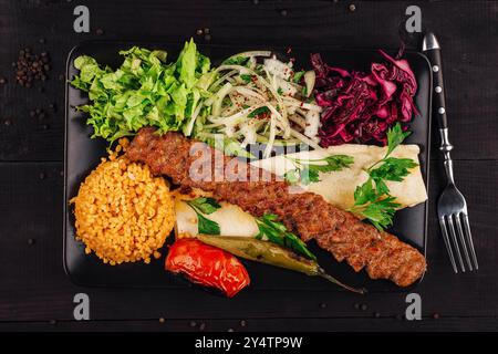 Kebab di adana della cucina turca con verdure fresche, bulgur e pane lavash su piatto nero su tavolo di legno Foto Stock