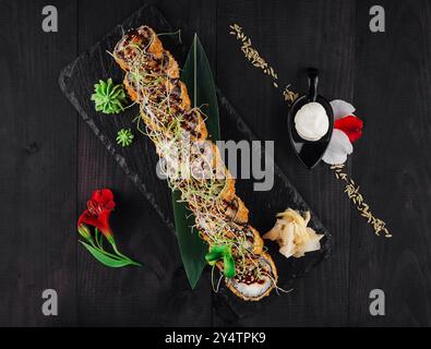 Il fresco rotolo di sushi tempura maki si trova su un piatto di ardesia nero decorato con fiori e semi di sesamo Foto Stock