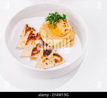 Piatto di hummus ricoperto di pinoli e olio d'oliva servito con pane pita e verdure fresche tagliate su sfondo bianco Foto Stock