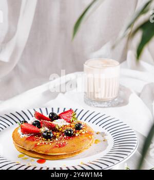 I pancake con panna montata, frutti di bosco e pistacchi si trovano su una tovaglia bianca vicino al cappuccino e alle foglie di palma Foto Stock