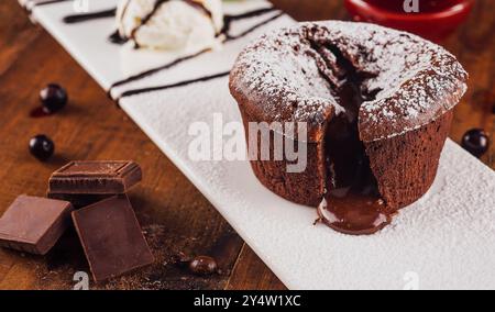 Torta lavica al cioccolato che trasuda cioccolato fuso su un piatto bianco, servita con una paletta di gelato alla vaniglia, frutti di bosco e pezzi di cioccolato Foto Stock
