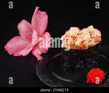 Sushi fresco con salsa piccante, decorato con caviale rosso, viene servito su un piatto di pietra nera accanto a un fiore rosa Foto Stock