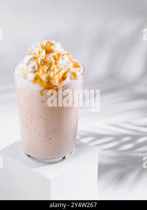 Il delizioso frullato di banana con panna montata e guarnizione al caramello si trova su un cubetto bianco su sfondo bianco con ombre Foto Stock