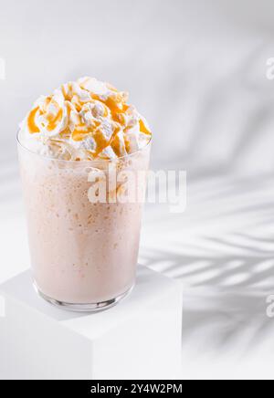 Bicchiere alto di milkshake al caramello con panna montata e salsa al caramello su sfondo bianco con ombre di foglie di palma Foto Stock