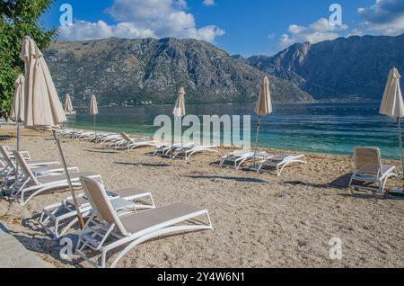 Vista idilliaca dalla spiaggia sabbiosa del villaggio di Stoliv, affacciata sulla baia di Cattaro con montagne e lettini vuoti, Montenegro, Europa Foto Stock