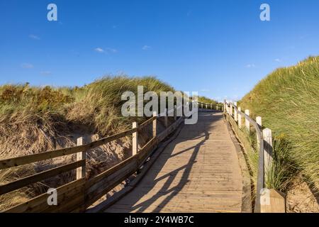 Guardando lungo un sentiero in legno sulla costa di Merseyside, con un cielo blu in alto e dune di sabbia ricoperte di marram su ogni lato Foto Stock