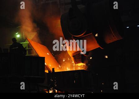 L'immagine cattura una scena drammatica all'interno di una fonderia o di un'acciaieria, mostrando il processo di versamento del metallo fuso da un grande mestolo in uno stampo o. Foto Stock