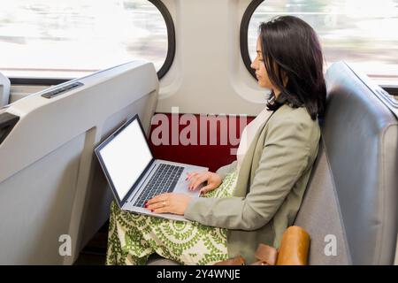 Una donna è seduta su un treno con un portatile aperto di fronte a lei. Indossa una giacca verde e una camicia bianca Foto Stock