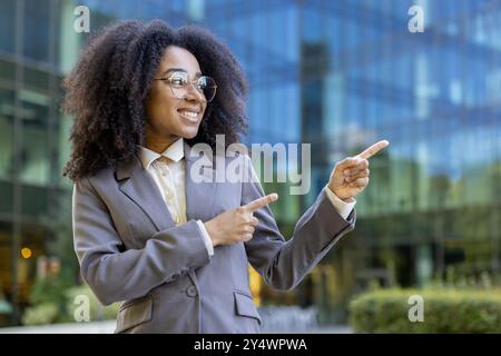 Donna d'affari sicura che sorride e punta con entrambe le mani mentre si trova all'esterno del moderno edificio di vetro. Donna professionista in abbigliamento formale rappresenta il successo e la positività Foto Stock