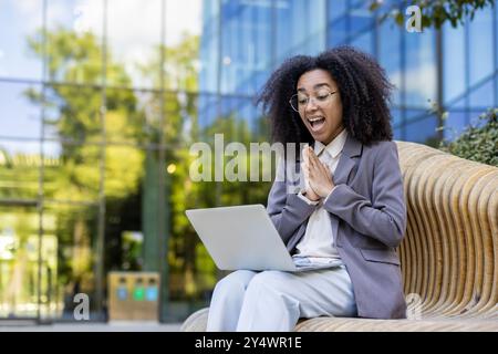 Donna d'affari ispanica seduta fuori dal moderno edificio di uffici usando un computer portatile. Sembra eccitata e gioiosa, vestita con abiti da lavoro. L'ambiente circostante mostra alberi verdi e facciata in vetro Foto Stock