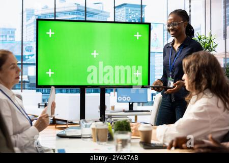 Diversi team di lavoro discutono le strategie aziendali con un grande schermo verde, riunendosi in una sala riunioni di fascia alta per esaminare le tendenze del mercato. Condividere nuove idee per la crescita e l'espansione del business. Foto Stock