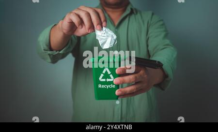 Uomo che indossa una camicia verde che tiene il cestino del riciclo e getta la carta nel cestino, concetto di smistamento dei rifiuti e sostenibilità, riciclo, vita sostenibile Foto Stock