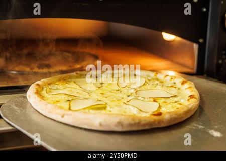 Una pizza appena sfornata con sottili fette di pera sopra, posta su un vassoio di metallo La pizza è in un forno a legna, con il vapore che sale sullo sfondo, Foto Stock