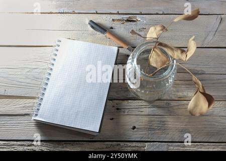 Mockup a spirale, penna e vaso in vetro con foglie marroni secche su un tavolo rustico realizzato con tavole di legno intemperie, ancora liv. Autunno e inverno Foto Stock