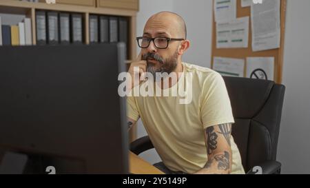 Bell'ispanico con barba e tatuaggi che lavora a una scrivania in un ufficio, molto concentrato sullo schermo di un computer in un ambiente di lavoro al chiuso Foto Stock
