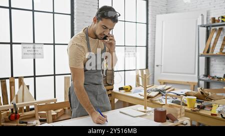 Uomo ispanico con barba in officina che usa il telefono mentre disegna piani su un banco da lavoro Foto Stock