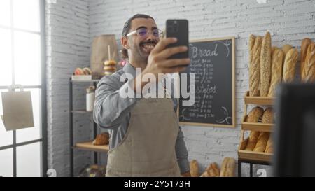 Giovane ispanico con i baffi sorridenti mentre si fa un selfie in una sala da forno al coperto, con pane fresco e pasticcini esposti sugli scaffali e un uomo Foto Stock
