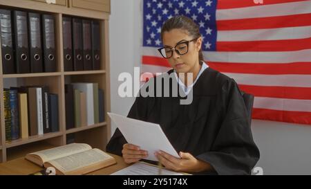 Giovane donna giudice che legge documenti in carica, con bandiera americana sullo sfondo, concentrandosi sul lavoro legale Foto Stock