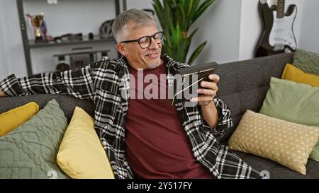 Un uomo dai capelli grigi si rilassa a casa su un divano, sorridendo mentre guarda un tablet in un salotto moderno. Foto Stock
