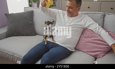 Uomo ispanico di mezza età seduto su un divano del soggiorno che gioca con due piccoli cani chihuahua. Foto Stock