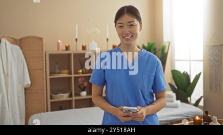 Terapeuta in uniforme blu sorridente in una serena sala termale con candele e piante, pronta a fornire un trattamento benessere Foto Stock