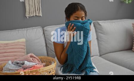 Giovane donna seduta al chiuso in un soggiorno che regge e puzza una coperta a maglia blu con un cestino di vestiti piegati accanto a lei su un divano grigio Foto Stock