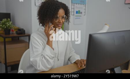 Una giovane donna afroamericana con i capelli ricci lavora in un ufficio, parla al telefono e usa un computer. Foto Stock
