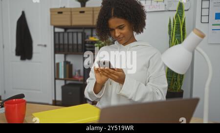 Giovane donna afroamericana con capelli ricci in una stanza d'ufficio, con cappuccio bianco, utilizzando uno smartphone, vicino a una scrivania con lampada, cartelle e menu à la Foto Stock