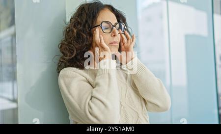Una donna ispanica matura con gli occhiali sta in casa, toccando le tempie come se fosse stressata o contemplando. Foto Stock