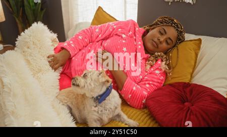 Donna afroamericana con trecce in pigiama rosa che dorme su un letto con un piccolo cane bianco in una camera accogliente. Foto Stock