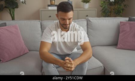 Bello, giovane ispanico con la barba seduto tranquillamente e con cura su un divano grigio in un salotto ben arredato di casa. Foto Stock