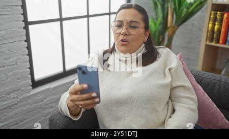 Donna ispanica che utilizza lo smartphone in un salotto accogliente, trasmettendo un senso di connessione e uno stile di vita moderno. Foto Stock