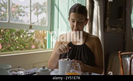 Donna che si gusta un caffè all'interno al tavolo del ristorante durante il giorno, la luce del sole scorre attraverso le finestre, circondata dal verde Foto Stock